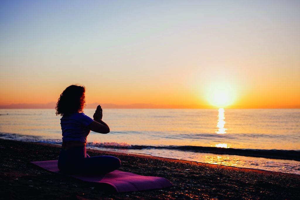 Carefree woman sitting in yoga mountain pose by the sea sunrise time. Finding inner peace spiritual healing lifestyle. Stretching in the morning for increasing energy.